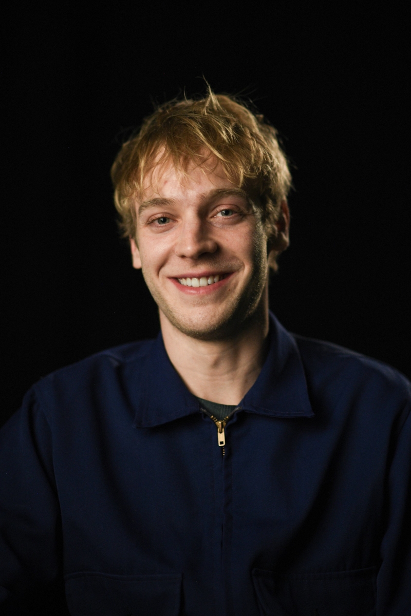 Headshot of a smiling caucasian male in late 20s, with blonde medium-length hair, wearing a blue jacket against a black backdrop.
