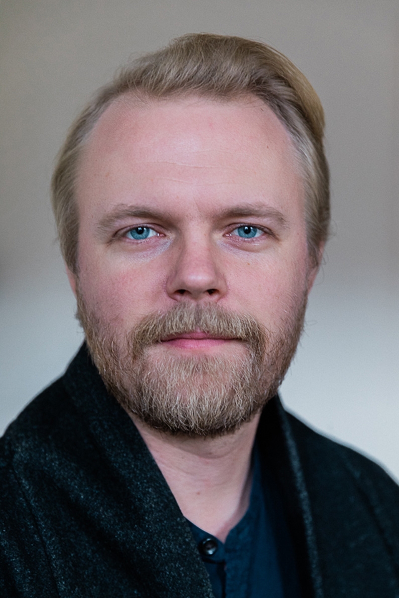 Headshot of an adult male with light skin tone, medium length blonde hair, beard wearing black wool cardigan over blue shirt, smiling slightly.