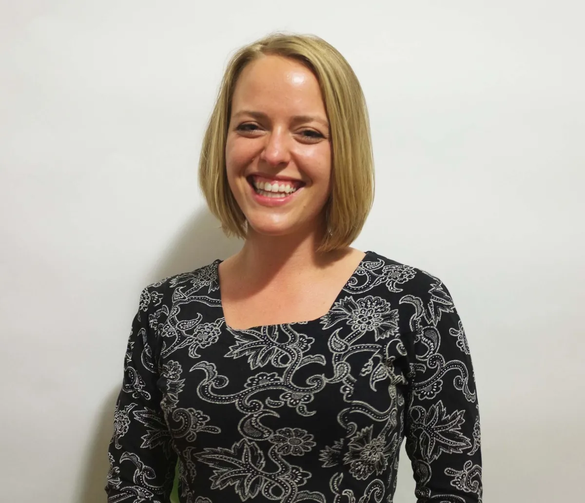 Headshot of a woman with light skin tone, blonde chin-length hair, wearing a black and white floral shirt. 