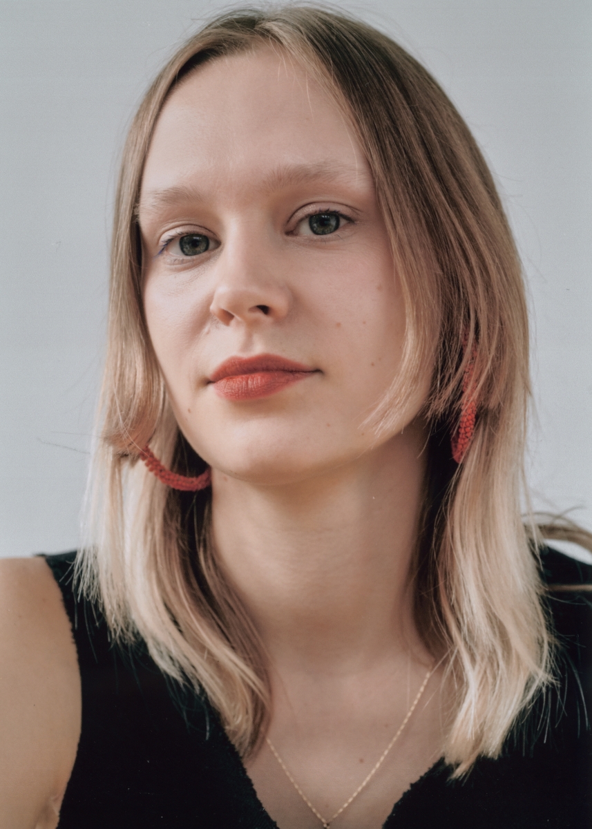 Headshot of a 30 year old White woman with light brown hair, wearing red earrings.