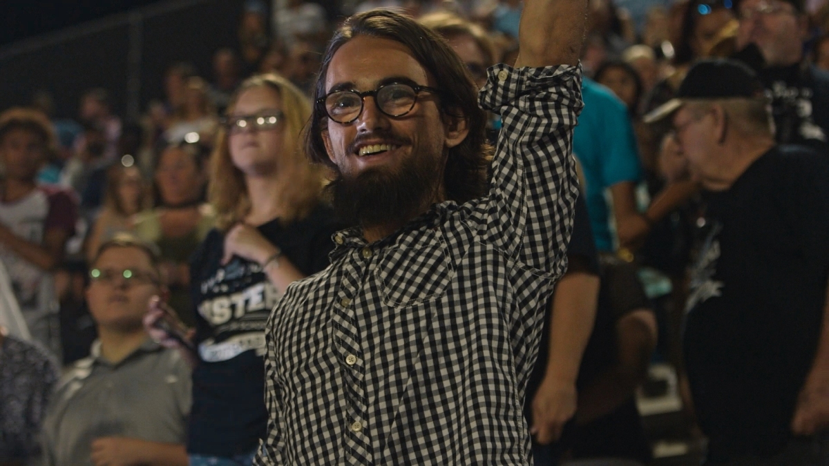 Film still from 'Your Friend, Memphis,' depicting a smiling young white man in a checkered shirt, raising his left arm, in a crowded stadium.