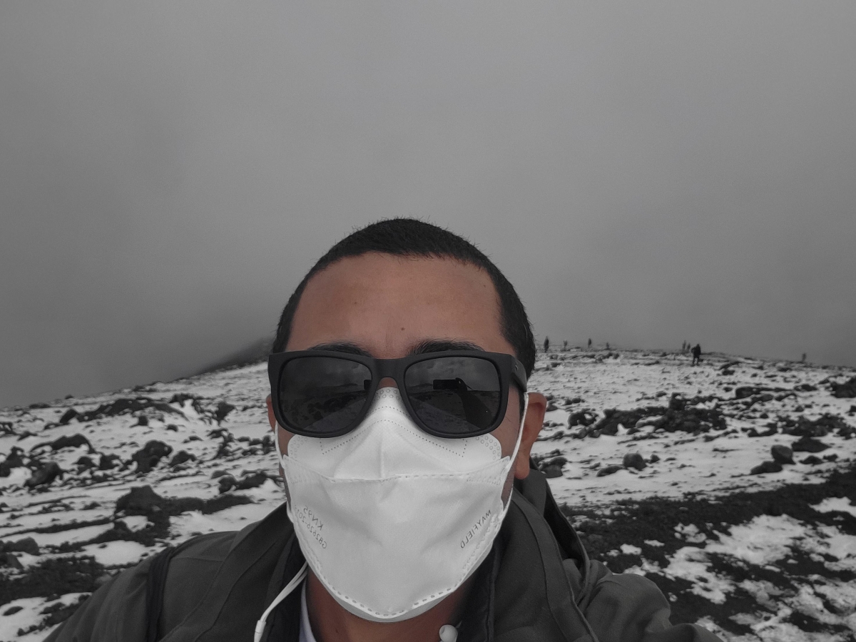Headshot of a mixed race middle-aged male in a mask and sunglasses, on top of a snowy hiking trail with smoke in the background.