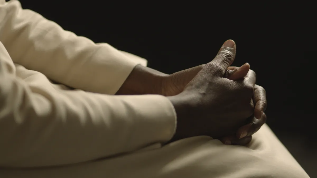 This picture shows a close-up of dark-skinned hands belonging to a Catholic nun dressed in a white robe against a black background. Her face can not be seen. The area in which the shot was taken is an interior illuminated with artificial light creating a high contrast.