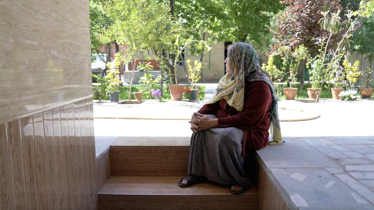 A female person, the main protagonist of the film, Hawa, sitting on the stairs of he appartement. She is looking away from the camera, watching a green garden. Hawa is wearing a colourful scarf covering her head, a red jacket and a grey dress.