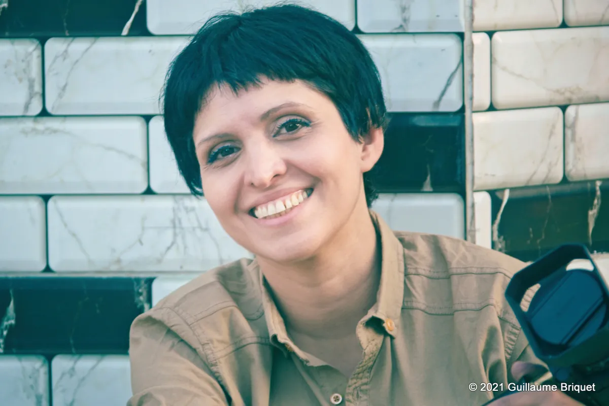 headshot of Maryam Ebrahimi wearing a brown shirt, sitting in front of a black and white brick wall holding a camera lens