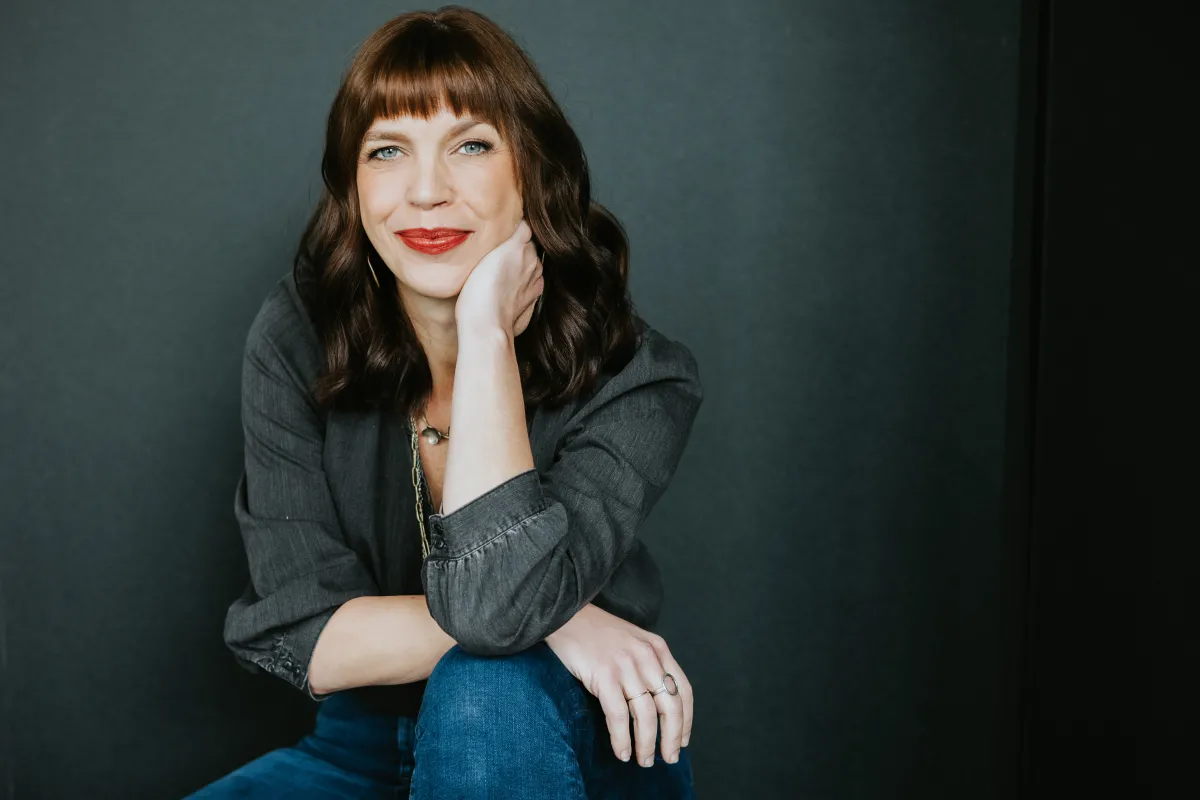 headshot of Sara Archambault wearing a dark gray top and blue jeans with red lipstick