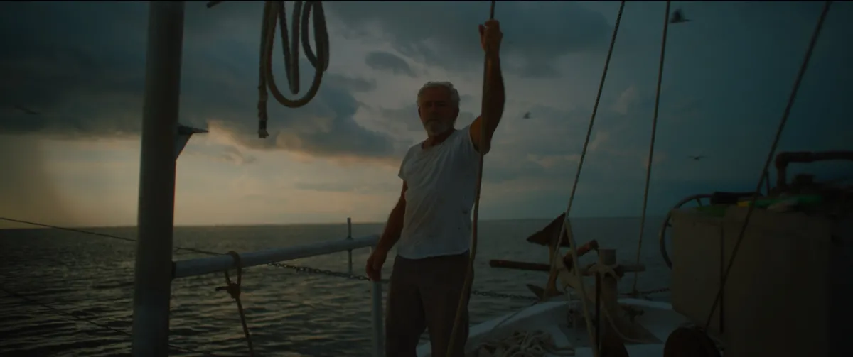 A nearly silhouetted silver-haired man stands on the bow of a boat, holding onto a rope above his head. Behind him is a cloudy dusk sky and an expanse of water.