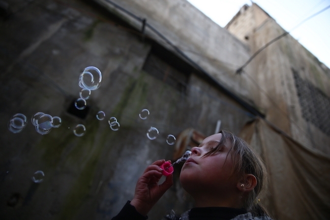 A young girl with hair in a ponytail blows bubbles in front of a large, dark building