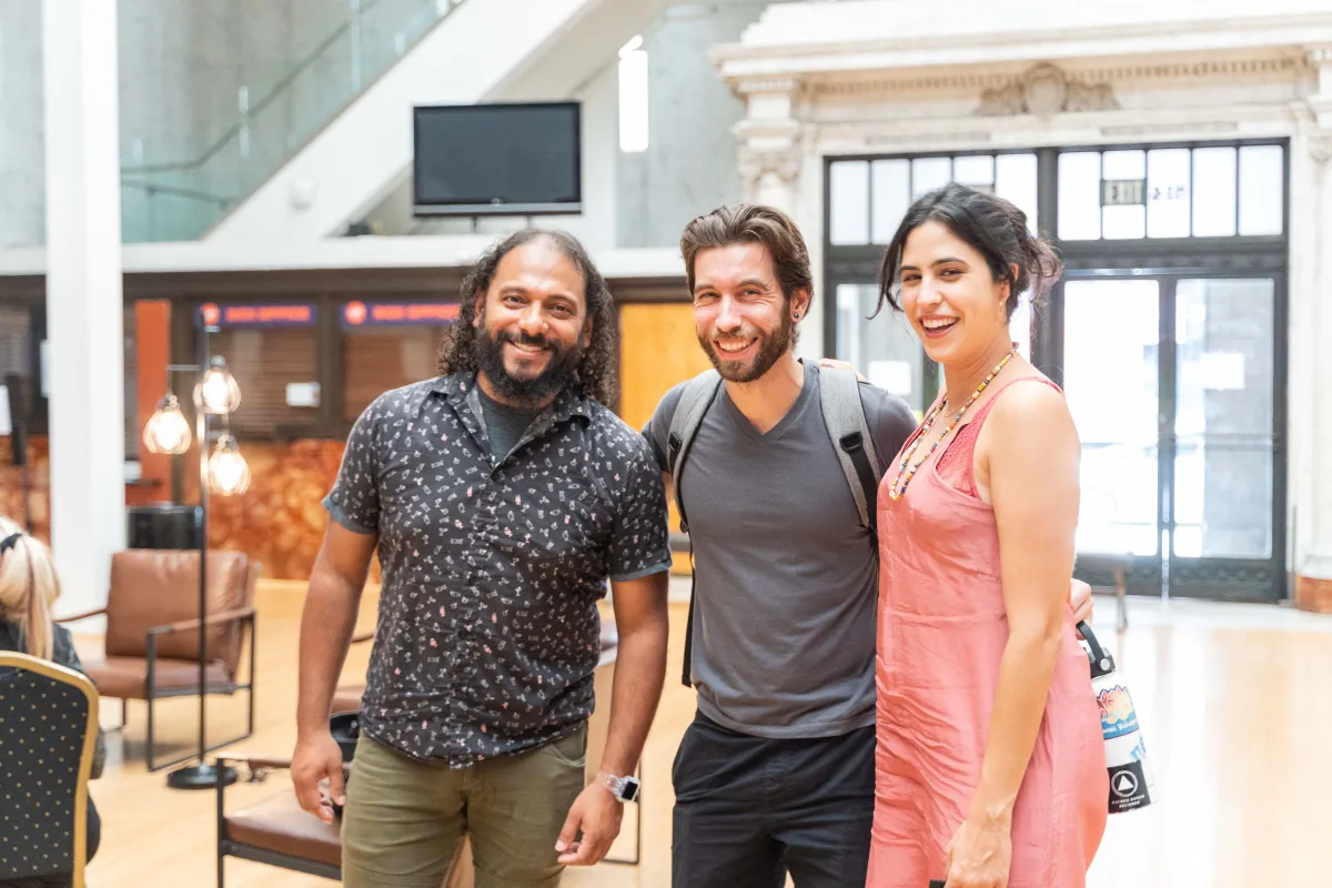 Three people standing in a brightly lit room, attending Getting Real '22. (left to right) Man is wearing a black and gray shirt and olive green pants, man in middle is wearing a backpack with a gray shirt and black pants, and woman on the right is wearing a pink tank top dress with her brown hair clipped in the back.