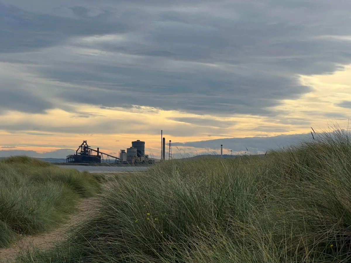 Landscape photo with tall green grass in the foreground with a sand path that leads to a river. Off in the distant background is a processing factory. The cloud filled sky with bits of sun poking through fills the rest of the landscape.