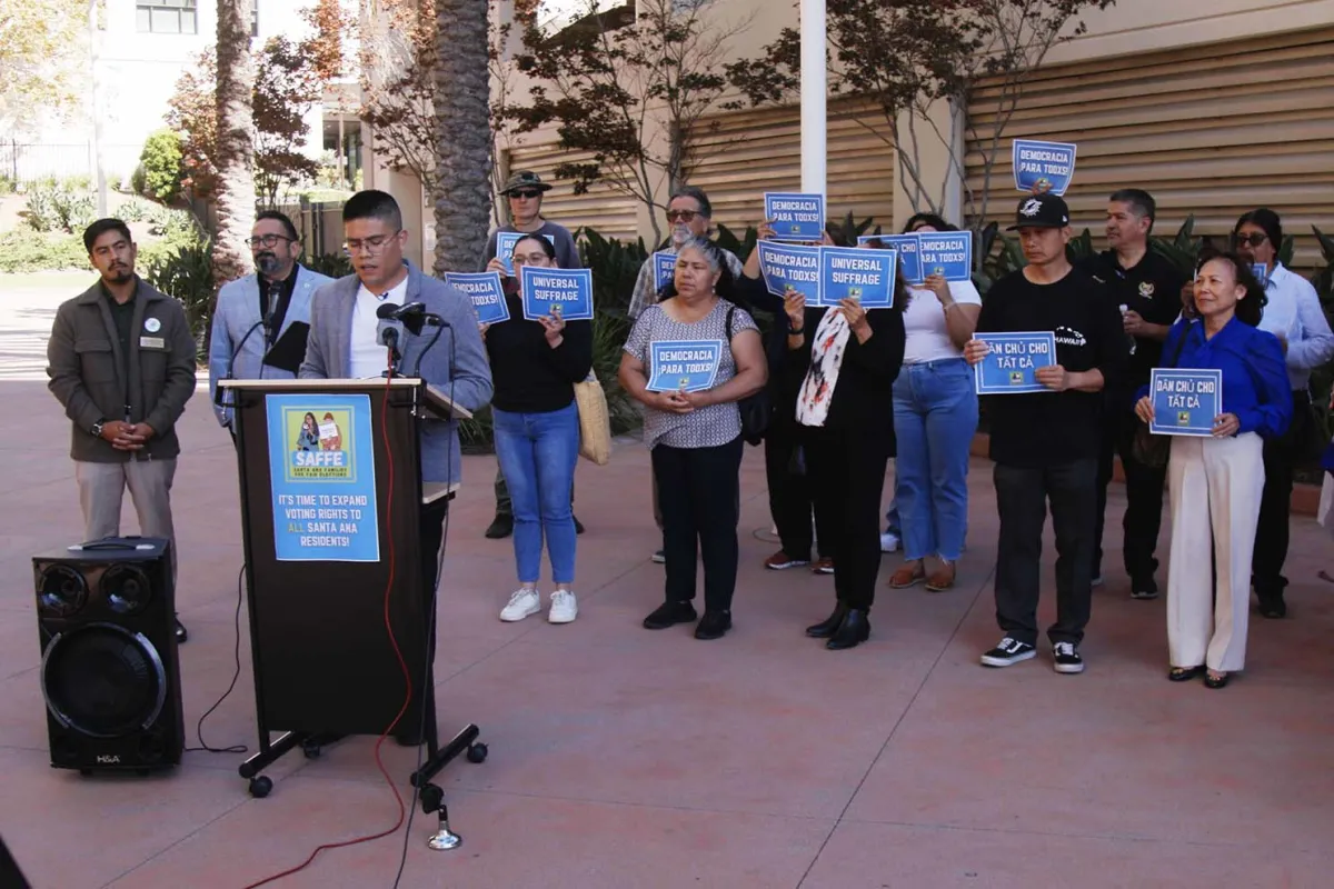 A group of protesters gathers behind a podium holding signs that say "Democracy for All" in English,Spanish and Vietnamese.