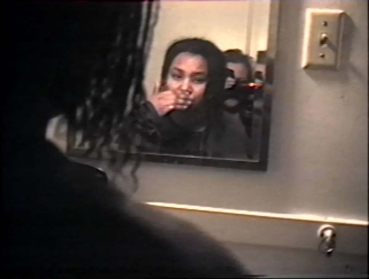 VHS still frame looking over the shoulder of someone in her early 20s with shoulder length dark hair into the mirror reflection where her hand is covering her mouth. A second person in her early 20s is visible in the mirror with a camcorder in front of her face.