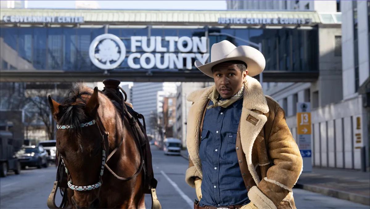 A man wearing a wide brim hat and a shearling jacket stands next to a brown horse in front of the Fulton County Courthouse