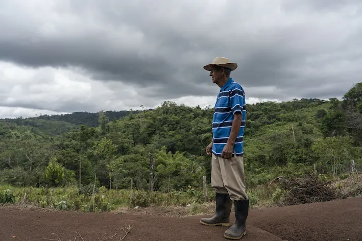 A man wearing a hat and rain boots stands in a hill, in front of some woods. There are dark clouds above