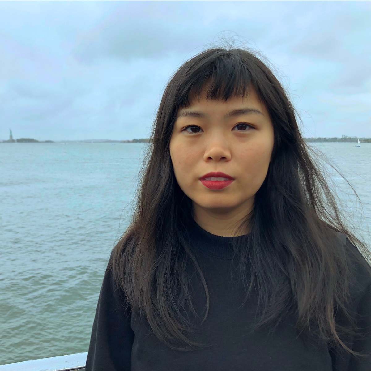 Yehui Zhao wearing a black shirt with long dark hair, bangs, and redish lipstick. A body of water and a clouds are in the back ground.