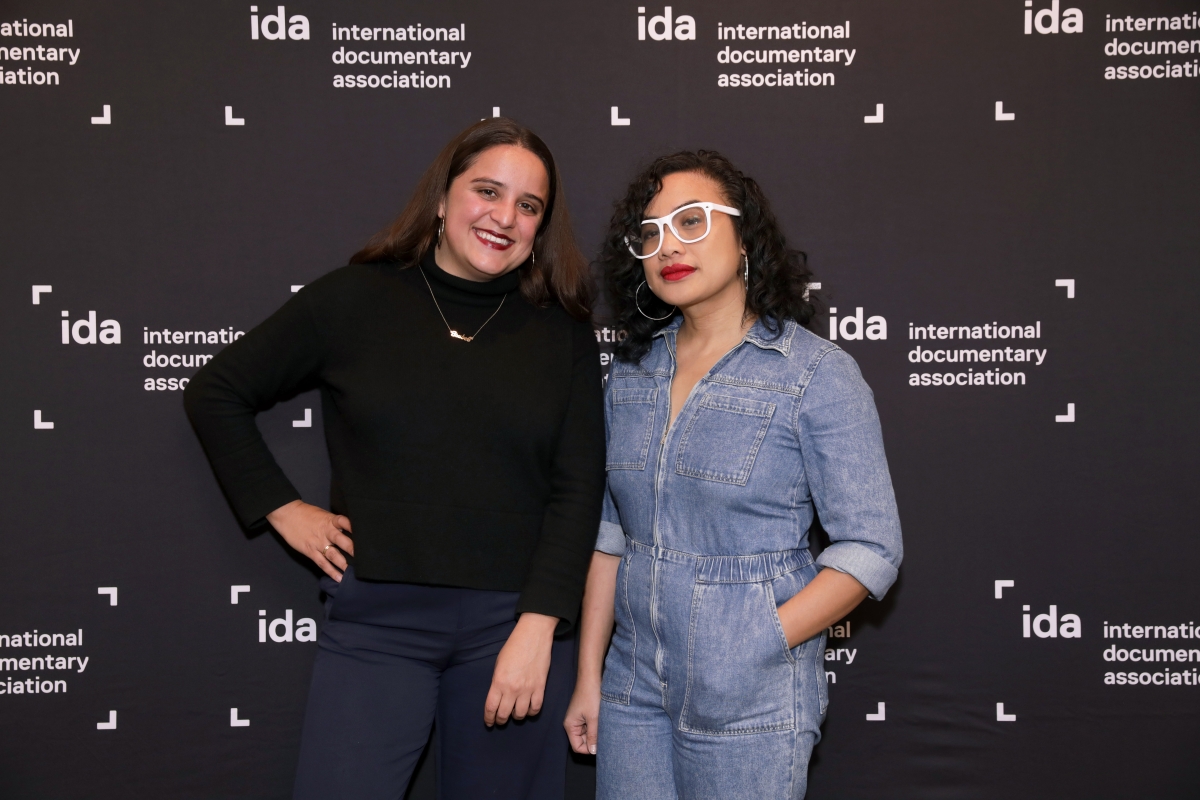 Two IDA members standing in front of a black step and repeat with white ida logos. Person on left is wearing all black, person on right is wearing a blue jean jumpsuit with glasses.