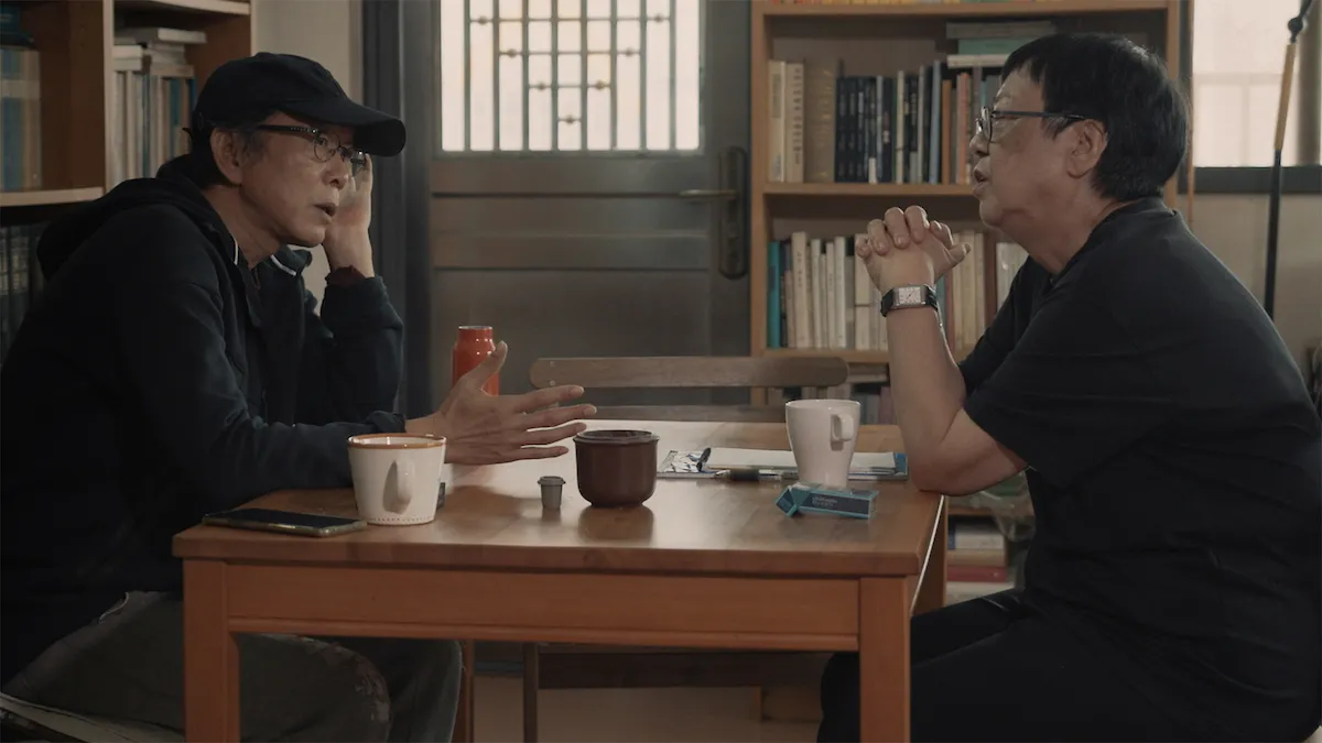 Two figures wearing dark shirts and glasses sit at a square table. They are surrounded by bookshelves, and are each drinking a mug of tea.