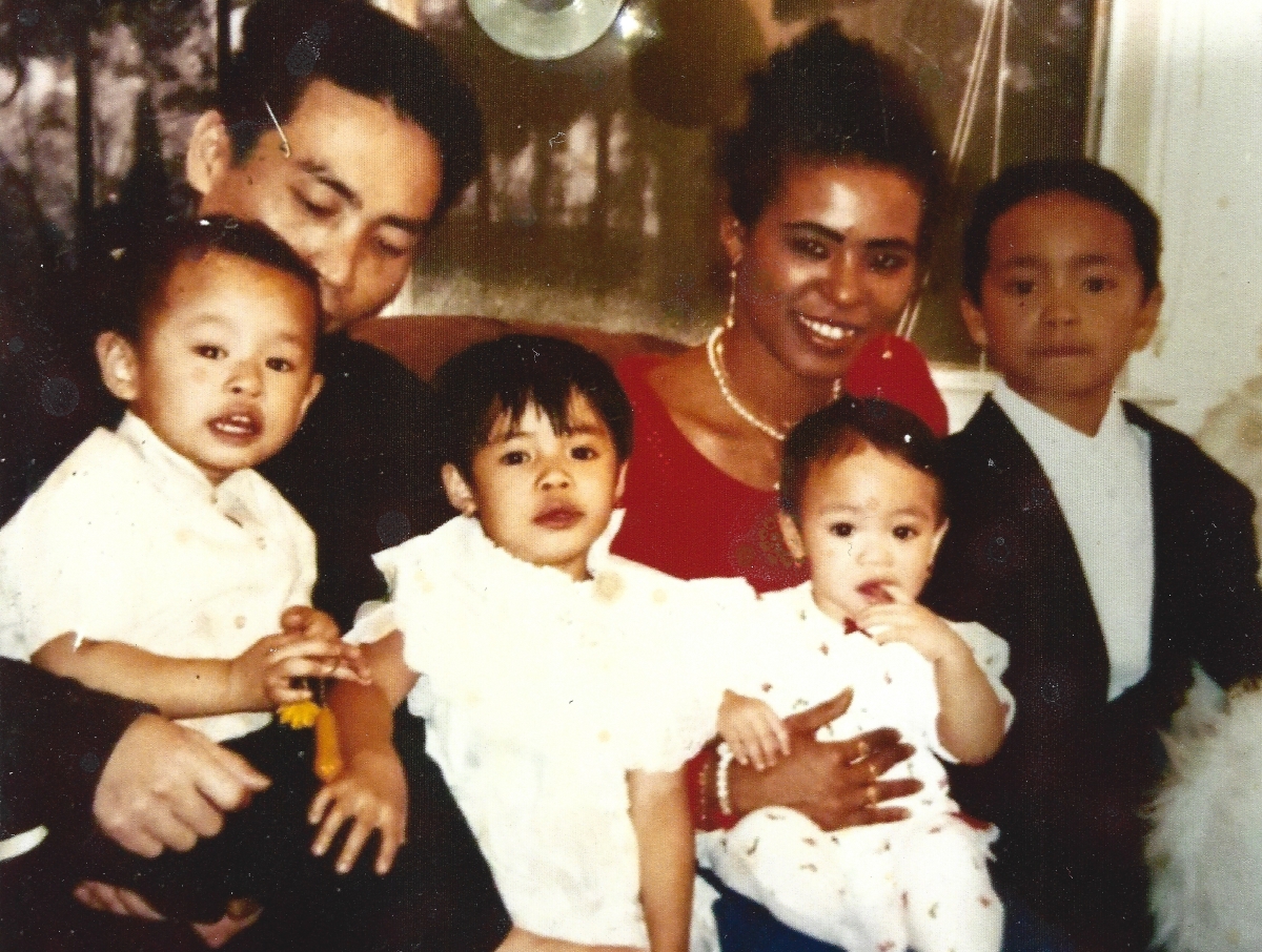 You see a blended Vietnamese and Amerasian family. There are 4 kids in the foreground. Three of them wearing White and one on the far left wearing a Black suit. The mother in the picture is wearing a red top with a pearl necklace.