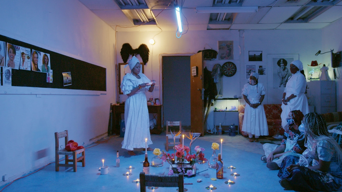 Still of a group of people in white dresses, conducting a ritual in a darkened studio.