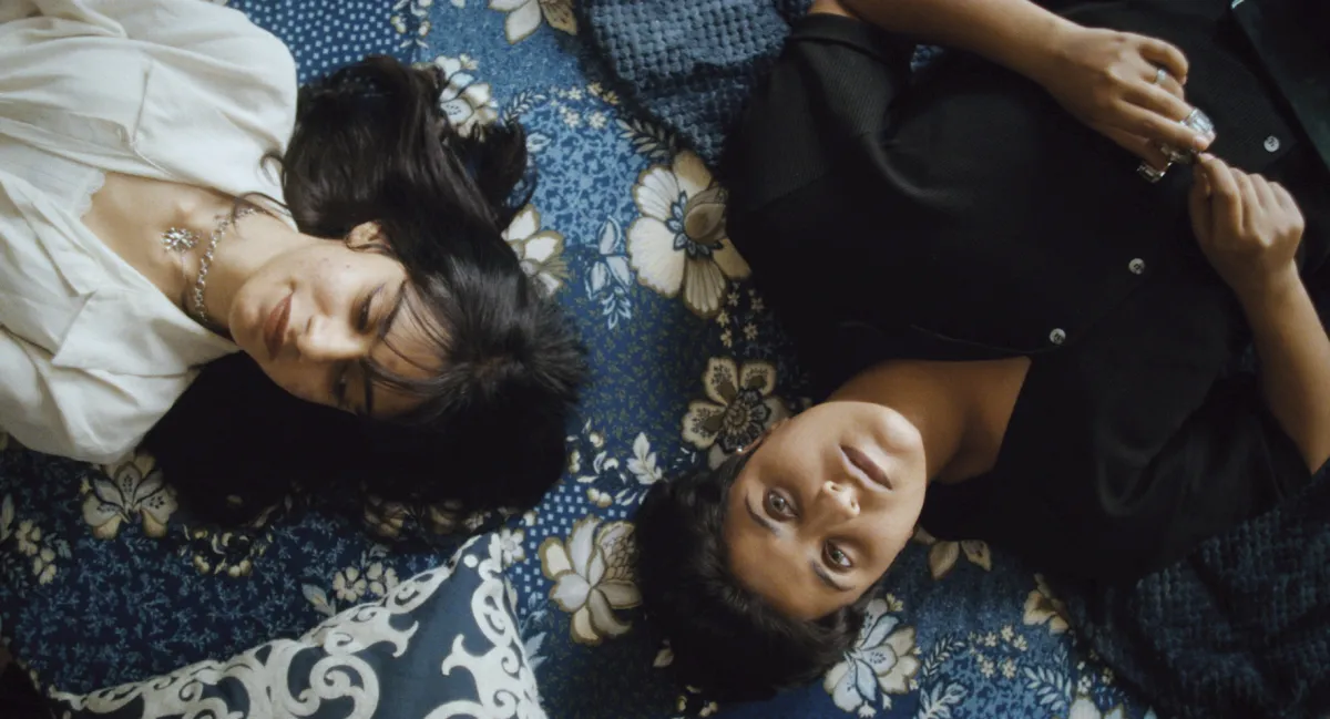 Two women lay on a bed with blue floral bedsheets.