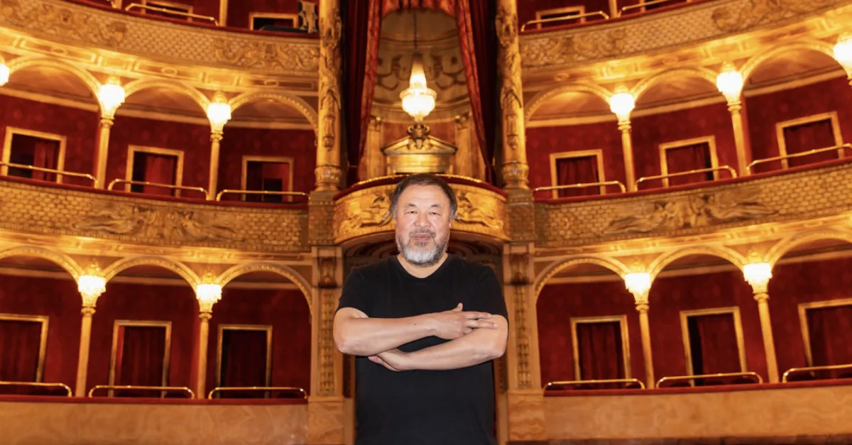 An Asian man wearing a black shirt stands with his arms crossed in front of the balcony seating of an opera house