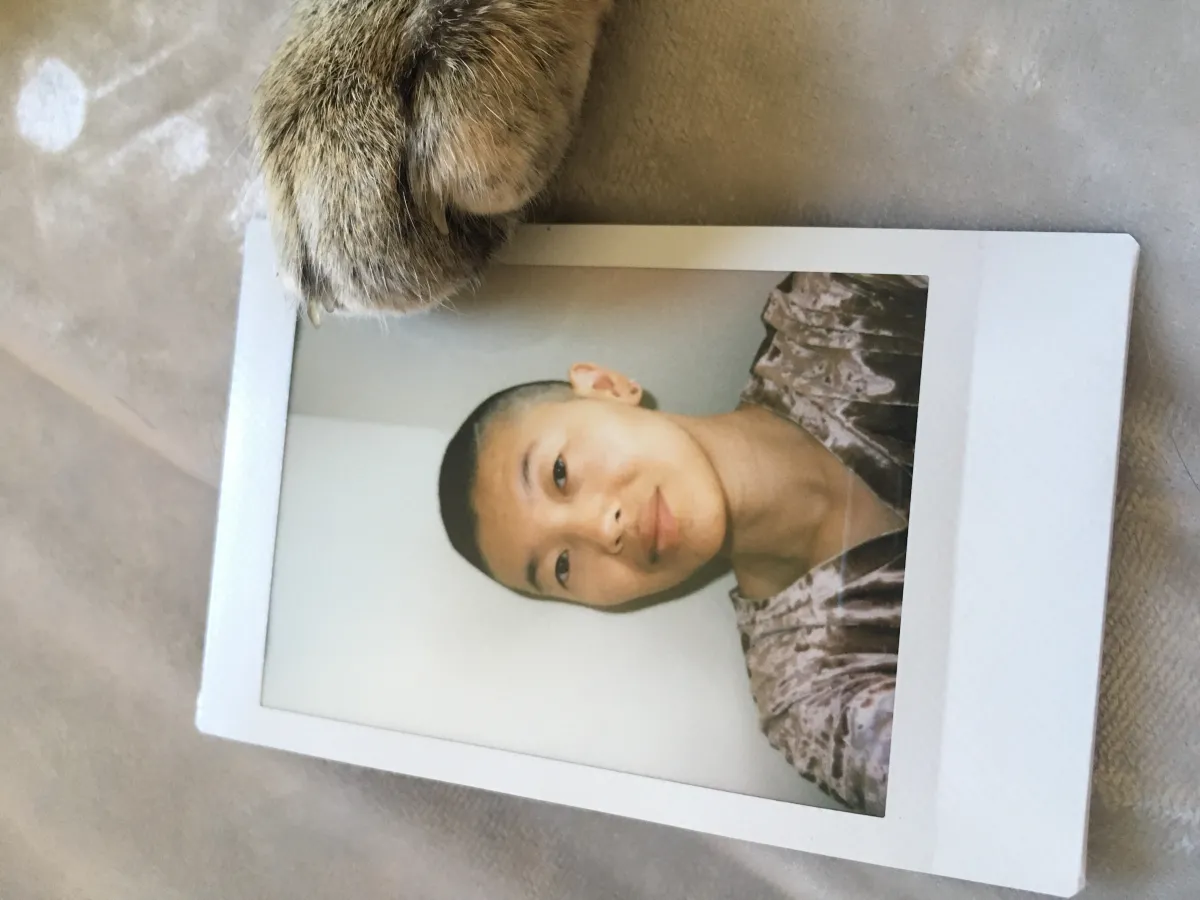 A photo of an androgynous person of Asian descent with a light smile at the camera. They have a dark shaved head and brown eyes and are wearing a pink velvet v-neck. The photo frames a photo print from a Polaroid camera; the print is lying flat on a silver texture, with a cat's paw gently lying on the top right corner of the print.