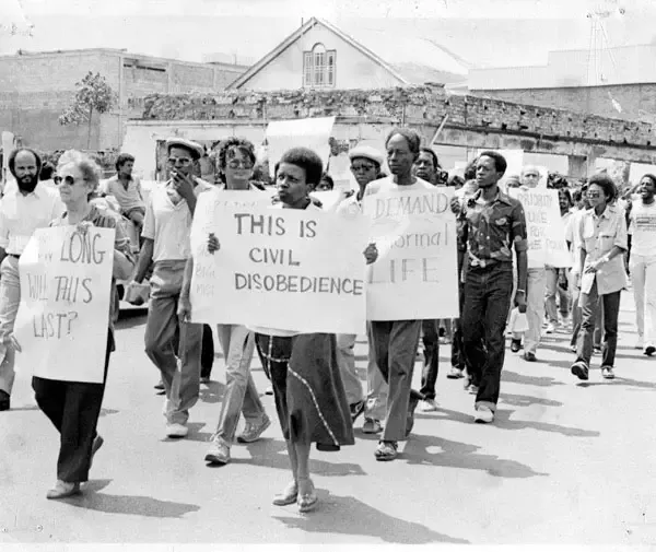 Black and white image of a group of people marching and holding protest signs. One sign says, "THIS IS CIVIL DISOBEDIENCE."