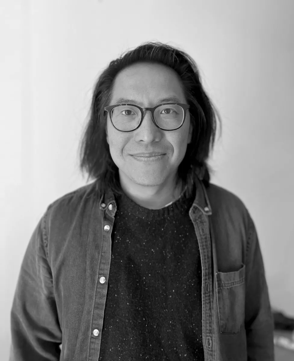 Black and White image of an Asian man. He is wearing black glasses and smiles at the camera. He has shoulder-length black hair and is wearing a dark sweater and button-down shirt.