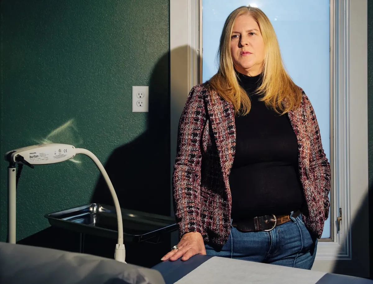 A middle-aged woman with long blonde hair, wearing a jacket and jeans, stands behind a medical exam chair.