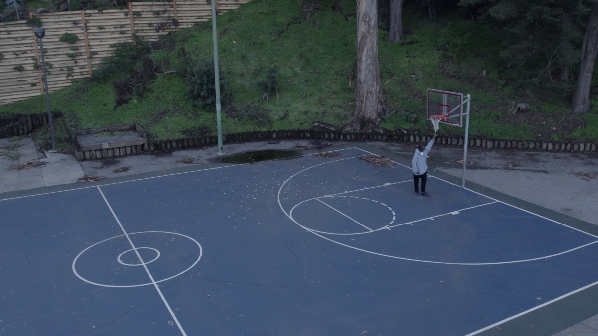 An African American man wearing a gray hoodie stands on an empty basketball court, reaching up to touch the basketball net