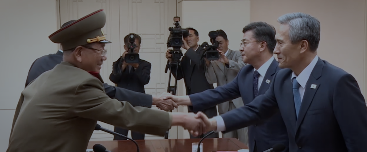 Two officials shake hands as a camera crew films them in the background.