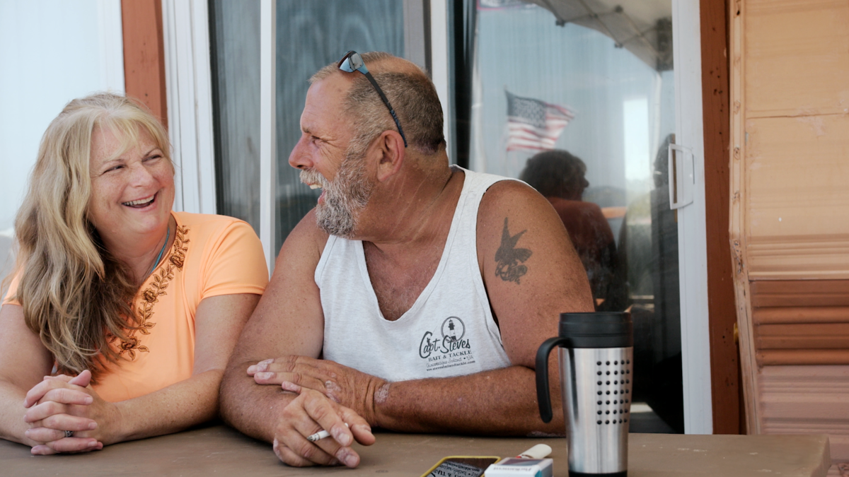 A middle–aged, white couple look at each other, laughing. The woman on the left is wearing an orange shirt and the man on the right is wearing sunglasses on his head and a white tank. He has a tattoo on his shoulder.