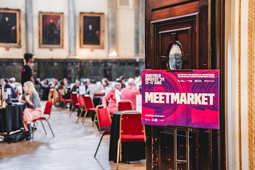 Doors open into a crowded ballroom with round tables, a sign for the Sheffield DocFest “MeetMarket” marks the event on the door.