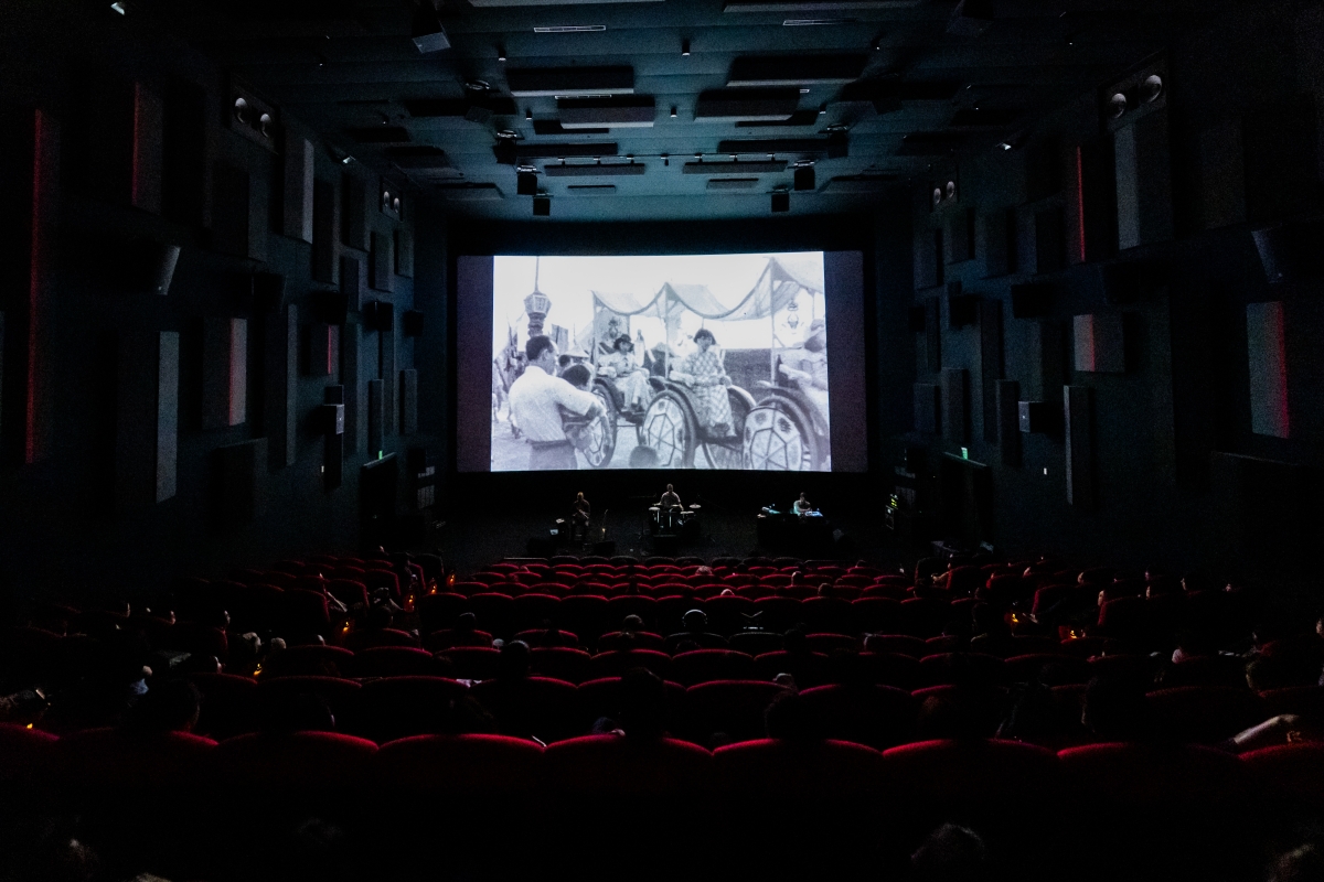 A large, dark theater screening “This Is Not a Film by Deng Nan-guang” with an experimental performance using drums and other instruments on the stage below the screen. 