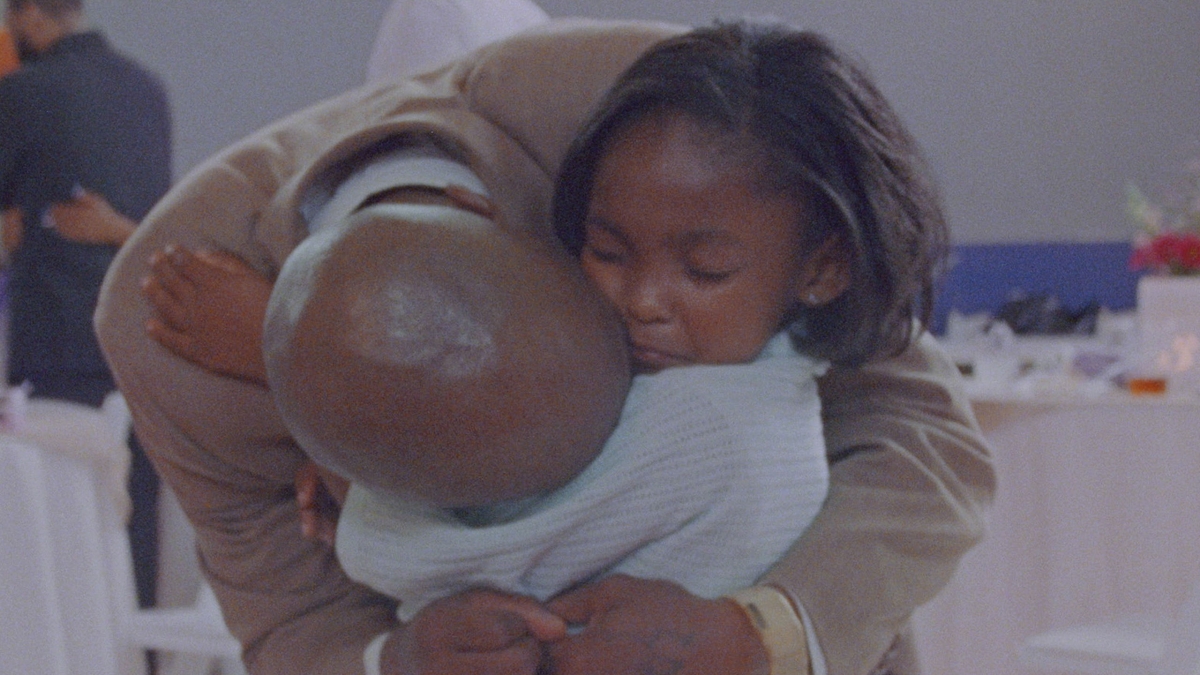 A bald man bends down to hug his daughter. The child is wearing a sad expression.