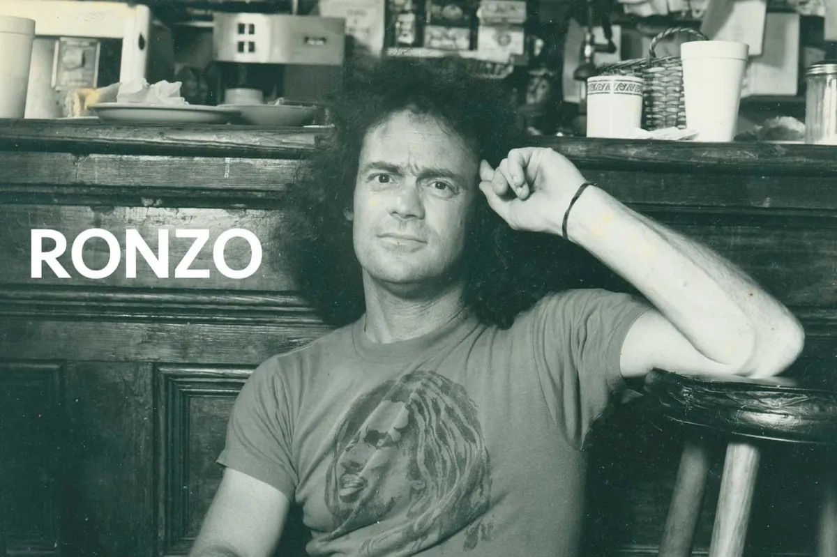 Black and white photograph of a man with dark, shoulder length hair leaning against a wooden cabinet