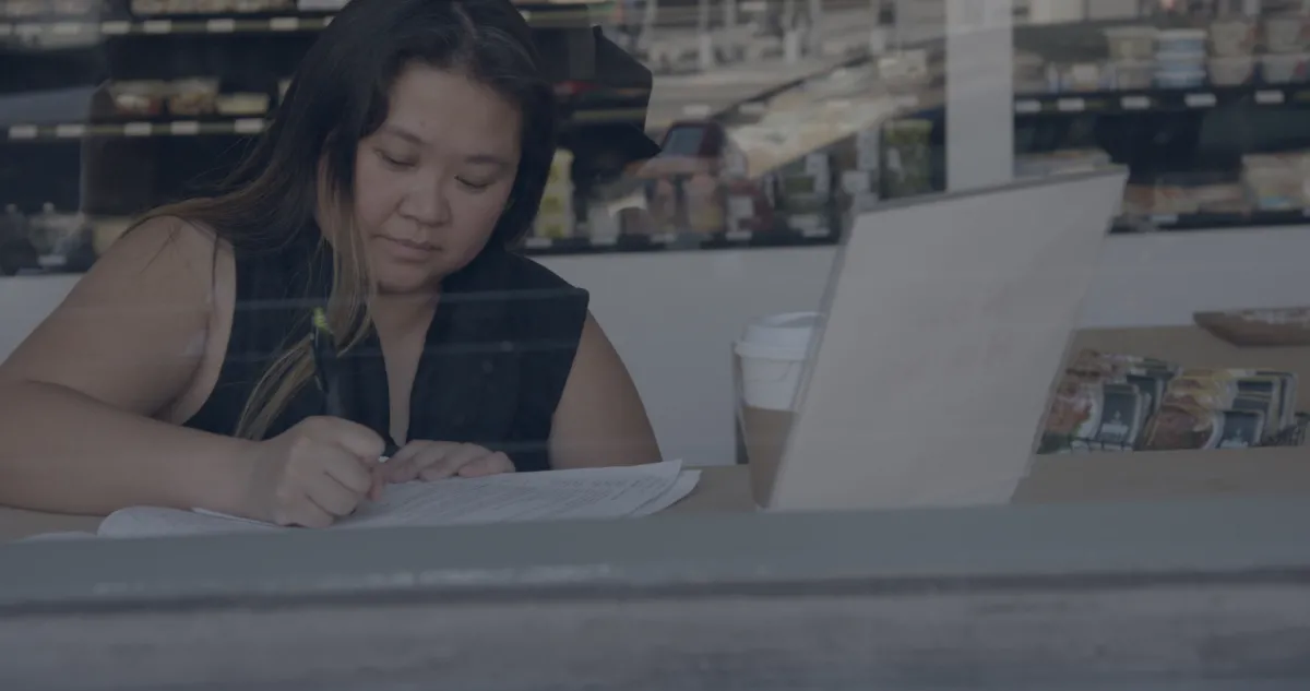 A woman with long hair and a dark top seen through a window writing in front of a laptop