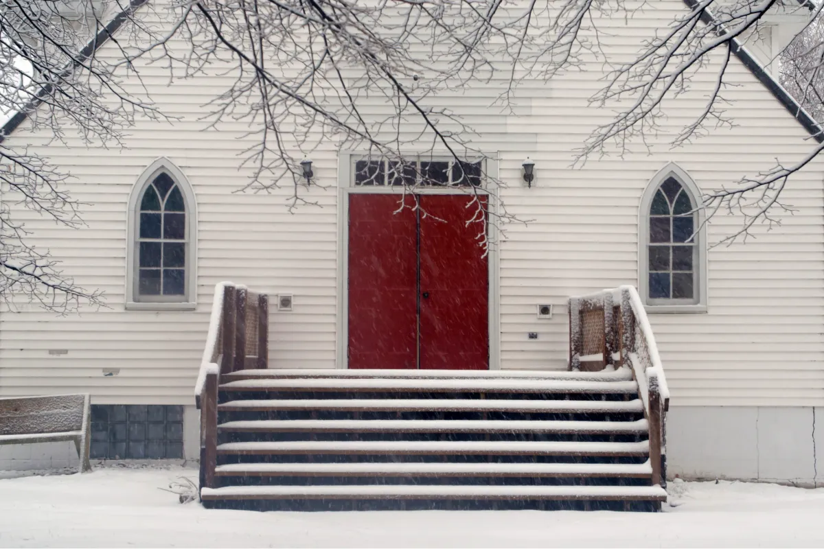 Snowy stairs lead up to a symmetrically-centered bright, red church door with a frosted church window on each side, and bare, snowy branches reaching toward the door from above
