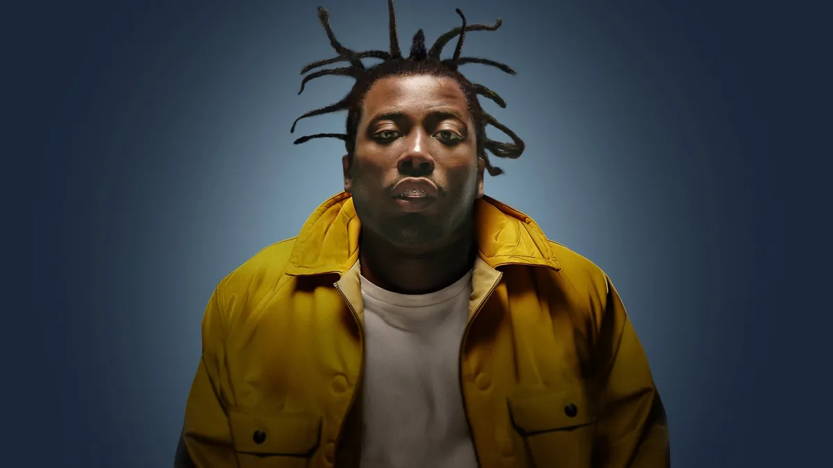 Photograph of a Black man with a yellow jacket, leaning into the camera.