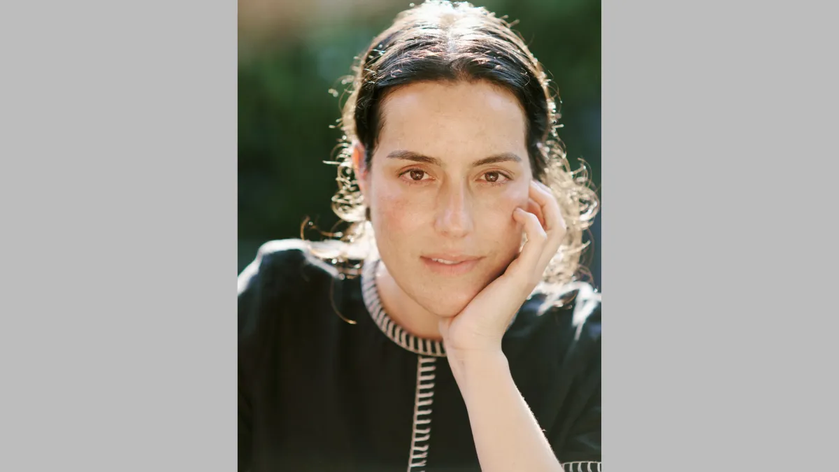 A woman with brown hair and wearing a black, colored shift leans her chin on one hand.