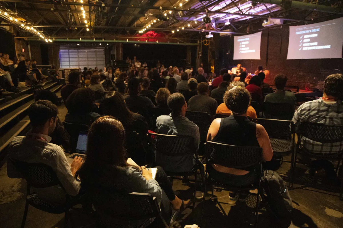 A crowded room of attendees sitting on fold-out chairs looks at a stage and presentation.