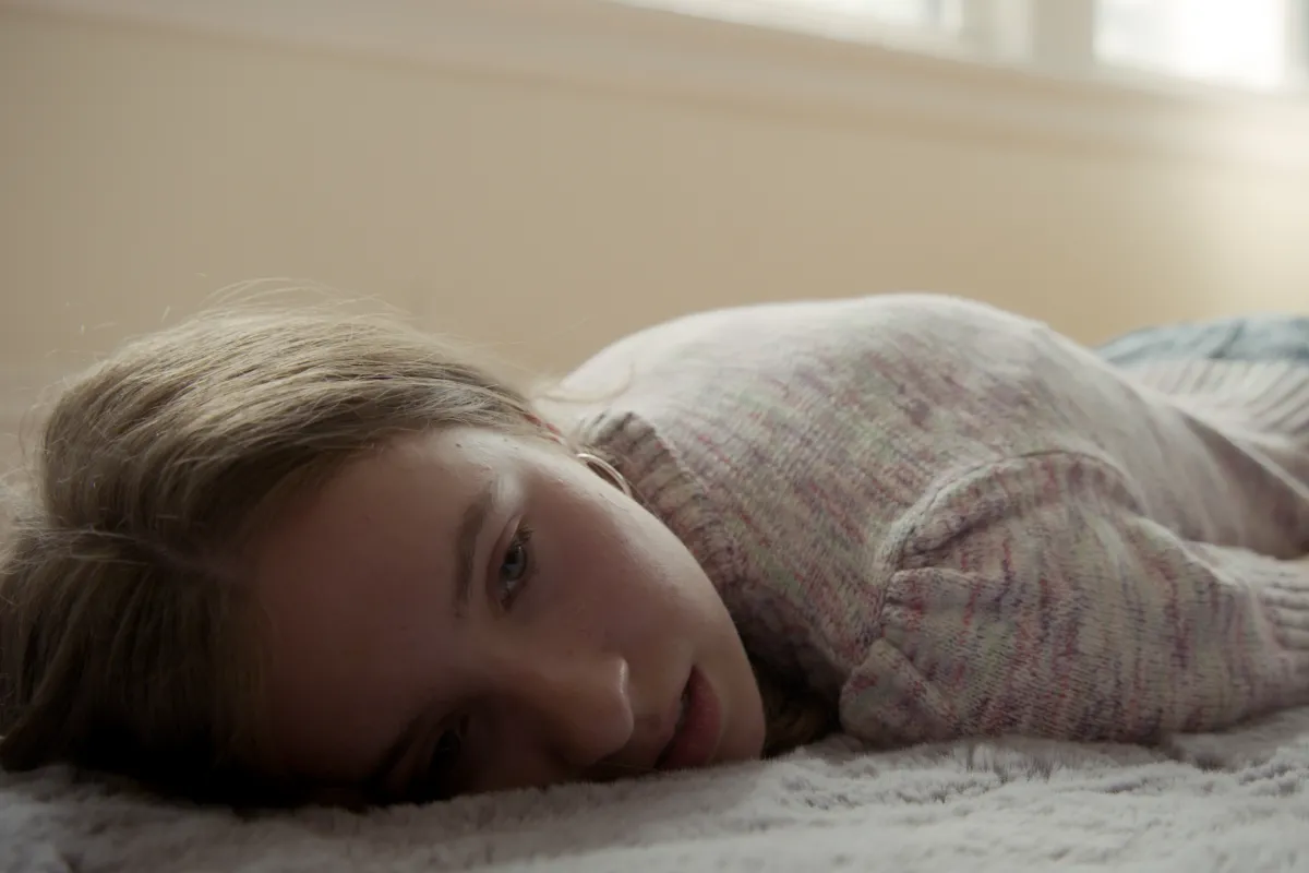 Close up of a teenage girl with light-brown hair and blue eyes, wearing a light-pink sweater and jeans, laying on her stomach on a bedroom floor with a noticeable, hunched-over spine, looking melancholily away from the camera.