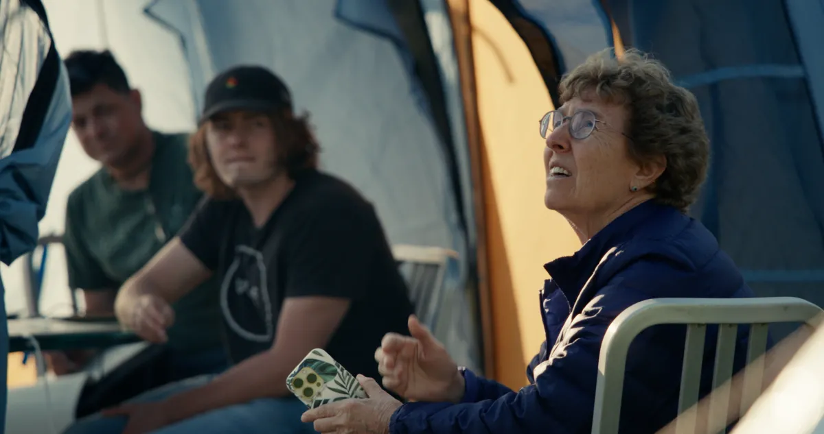 During golden hour, an older bespectacled astronomer looks up anxiously to the sky. Her younger colleagues, at a distance in a tent behind her, look to her. All are concerned.  