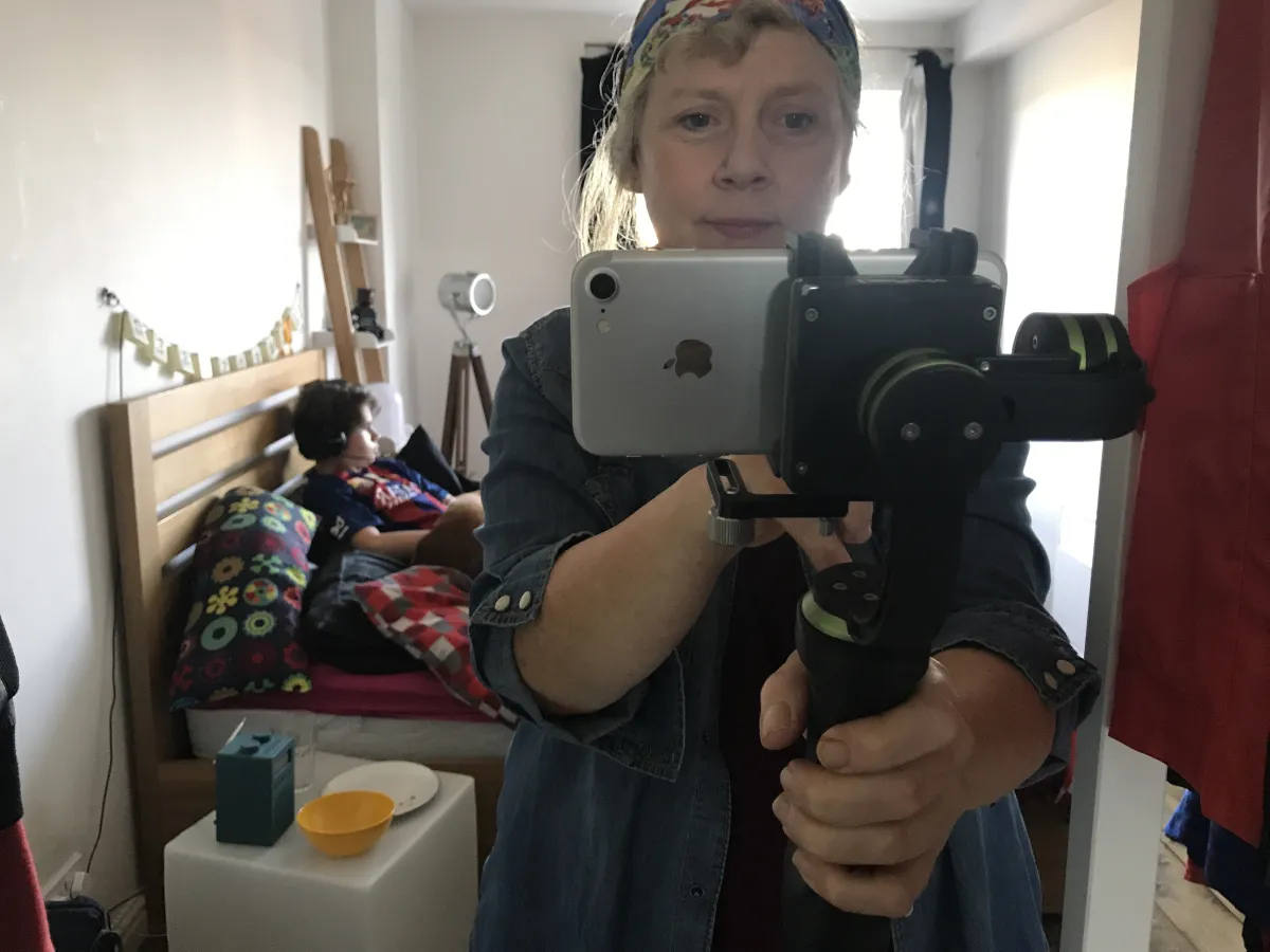 A white woman films herself with an iPhone. Behind her, on a bed, is her teenaged son.