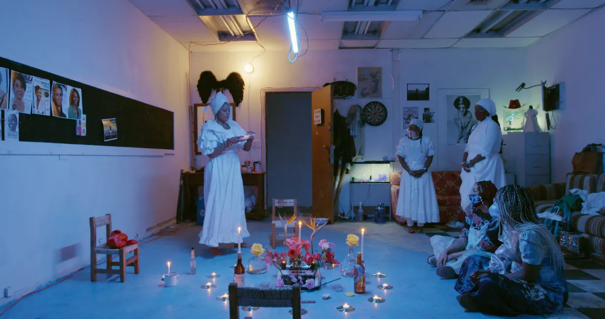 blue light colored a room of three balck women dressed in all white conducting a ritual while a group of people watched 