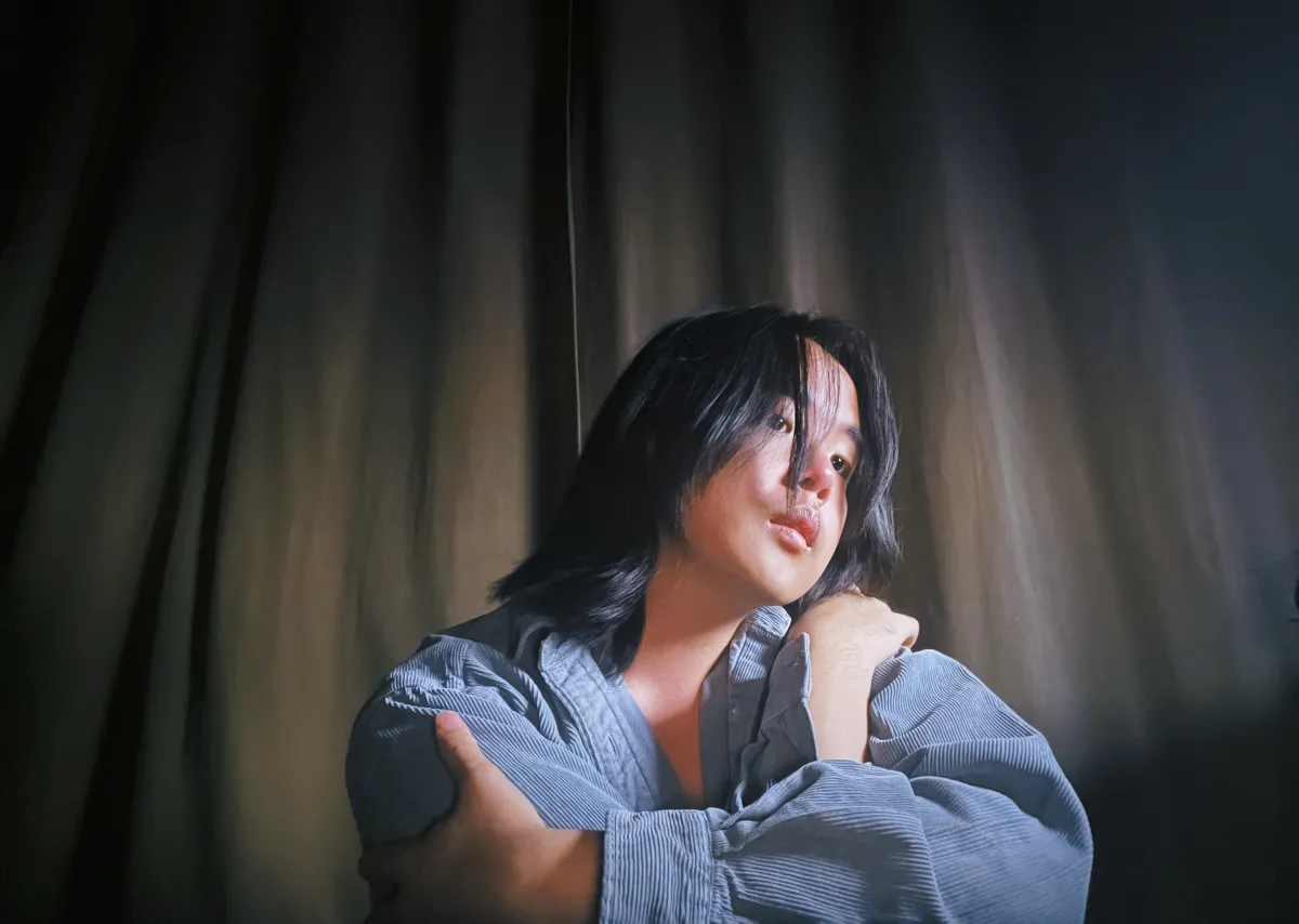 Los Angeles-based composer Lucy Liyou sits in front of a curtain, wearing a long-sleeved blue shirt.