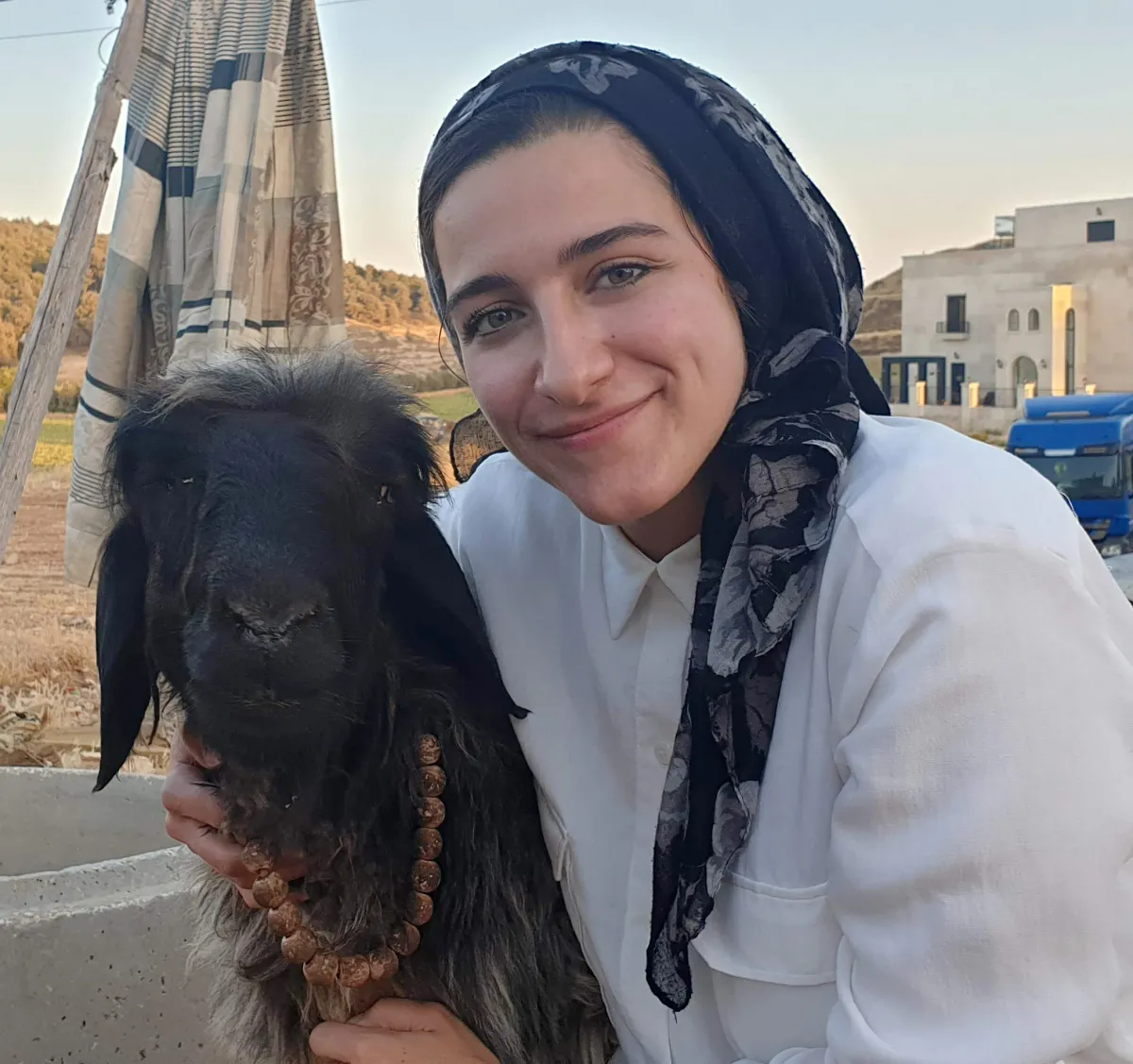 A person with a head scarf hugs and goat and smiles.