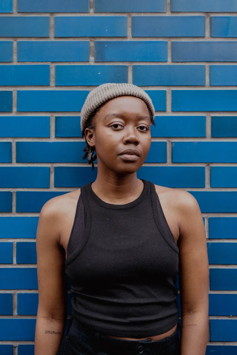 Headshot of Jameisha. Brown skin person wearing a black tank top and gray beanie, standing against a blue brick wall.