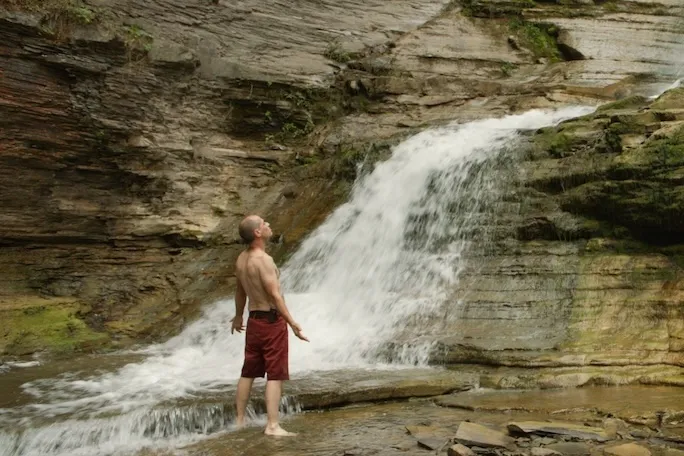 A shirtless man stands in front of a waterfall with his arms spread