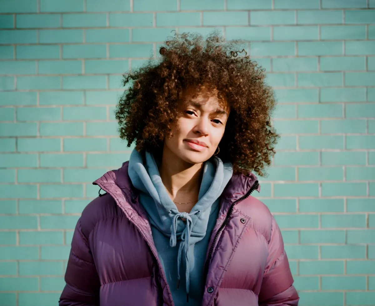 Headshot of a black woman with an afro and a puffer jacket in front of a green brick wall